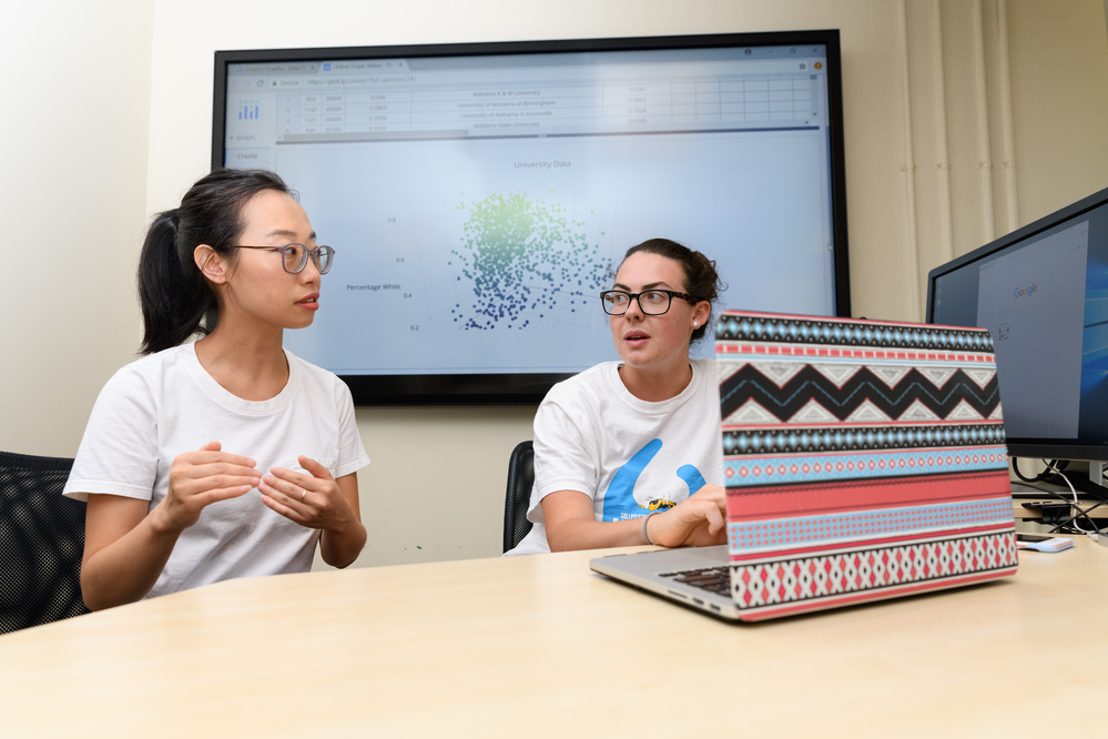 Tech employee talking while sat at a table with a laptop and screen behind them