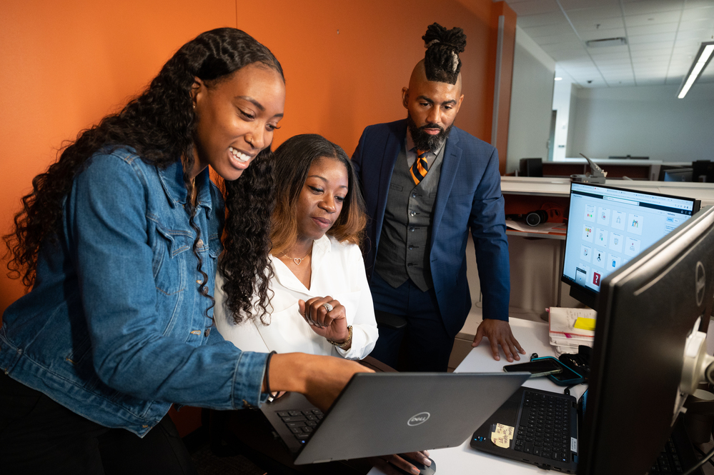 3 Tech employees looking at a laptop while standing around a desk